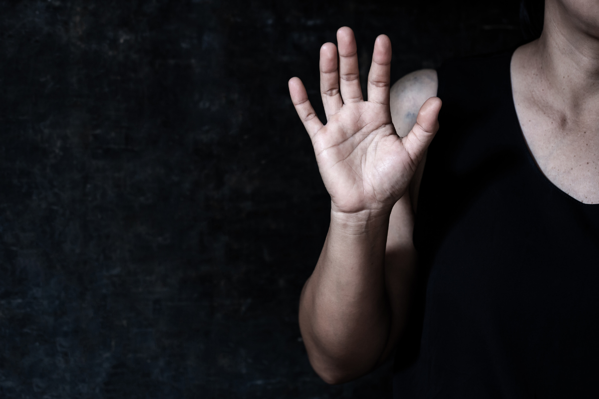 A person in a black tank top holding up their open palm in a stop gesture against a dark, textured background.