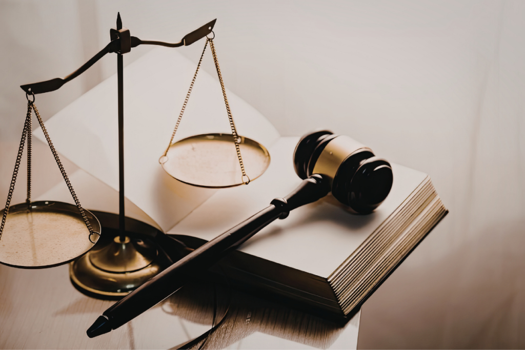 A wooden judge's gavel resting on an open law book next to a brass scale of justice on a wooden table.
