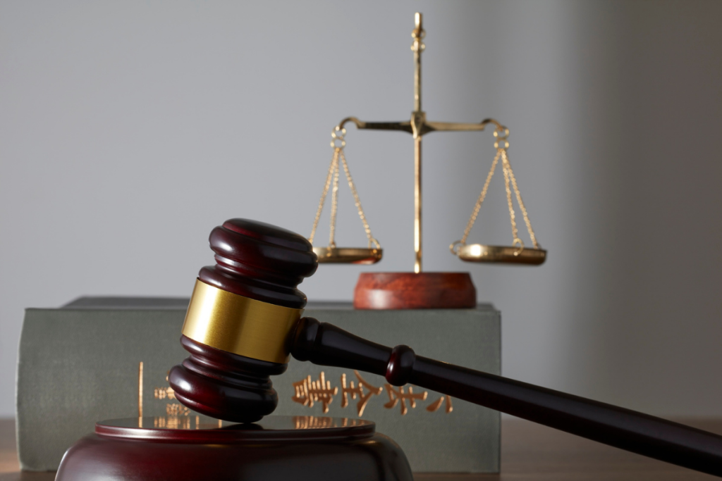 A wooden judge's gavel resting on a sound block in front of a law book and a golden scale of justice.
