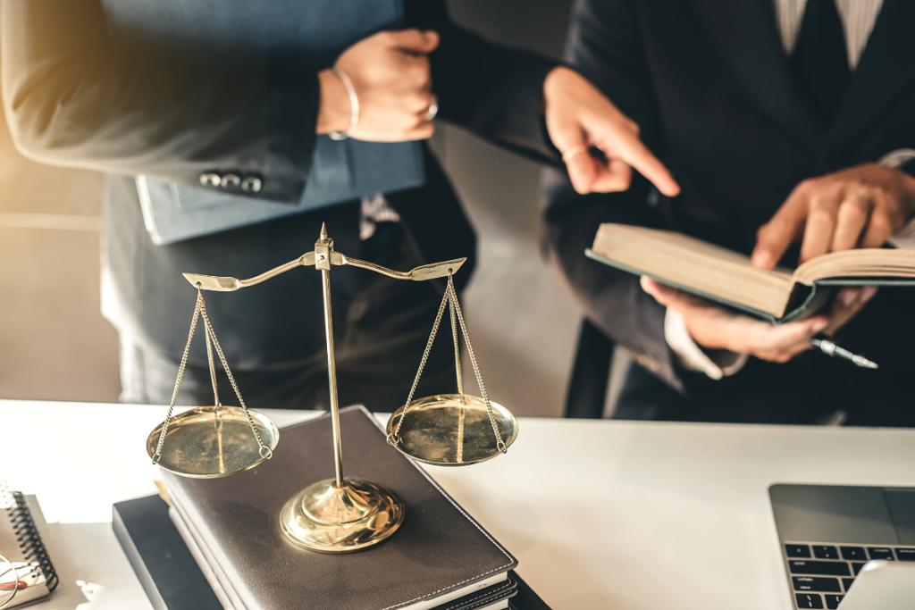 A brass scale of justice sits on law books while two lawyers in suits consult a book in a professional office.
