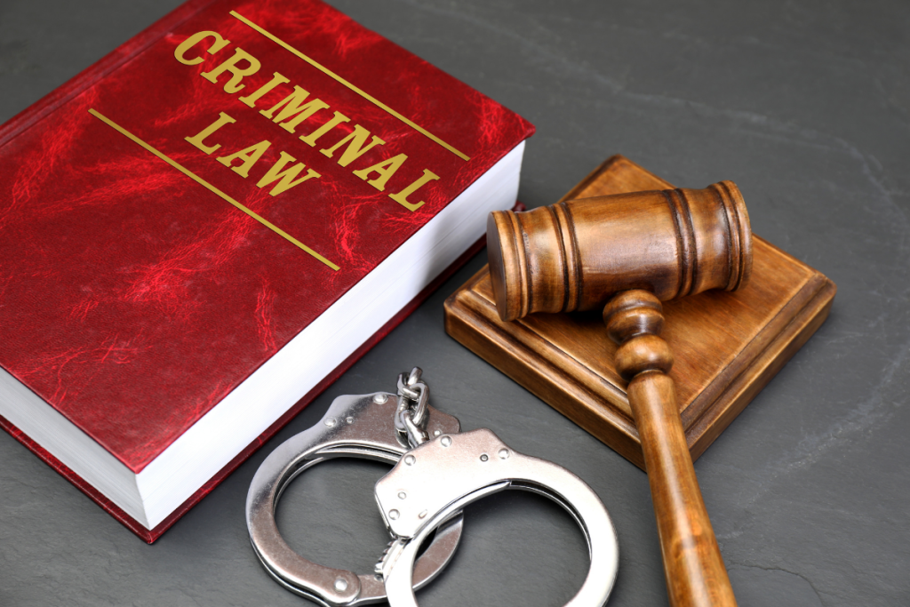 A red Criminal Law book, a wooden judge's gavel, and metal handcuffs on a dark slate background.
