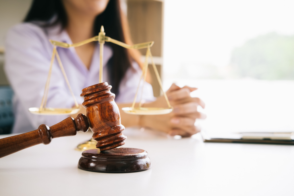 A wooden judge's gavel resting on a desk in front of a blurred lawyer with a gold scale of justice.