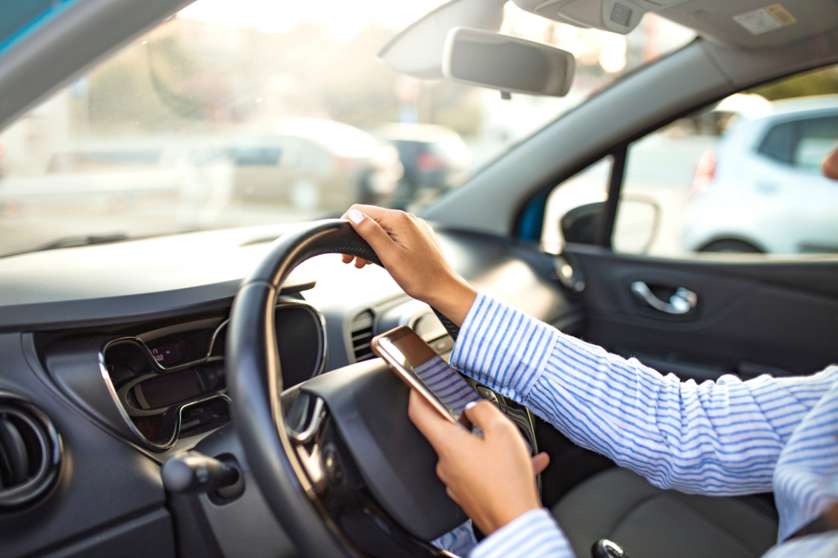 A driver holding a smartphone while steering a car, highlighting the danger of distracted driving.