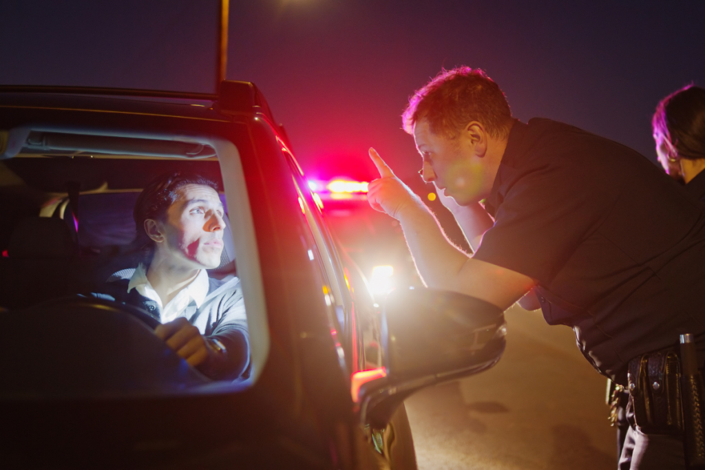 A police officer points at a driver during a nighttime traffic stop with emergency lights flashing in the background.